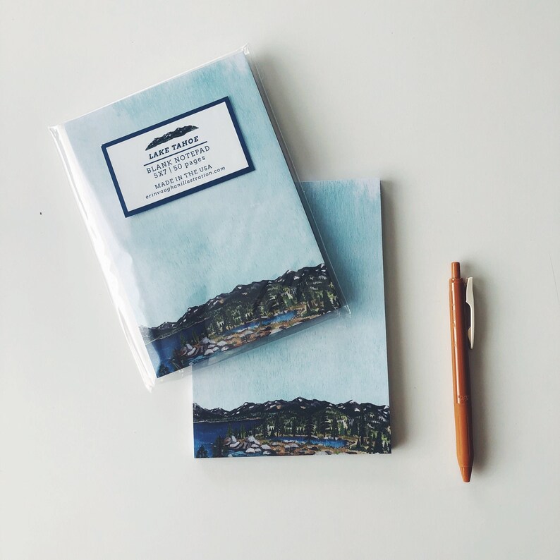 May include: Two Lake Tahoe blank notepads, 5x7 inches, with a watercolor landscape design of mountains and a lake. A brown pen rests beside the notepads. The notepads are packaged in clear plastic.