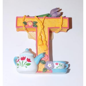 Op de afbeelding: Een decoratieve letter "T" in geel met roze randen, versierd met een paarse tulp, een theepot en een theekopje. De theepot en het theekopje zijn wit met rode bloemmotieven. Een geel touw met kwastjes voegt details toe.