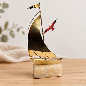 Pode incluir: Uma estatueta decorativa de veleiro feita de metal dourado polido, com uma vela curva, uma pequena bandeira no topo do mastro e um detalhe de pássaro vermelho. O barco está montado sobre uma base retangular de pedra texturizada em tom off-white.