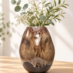 May include: A rounded glass vase featuring a glossy finish with a blend of brown, tan, and cream tones that transition into a solid dark brown base. The vase contains a bouquet of green eucalyptus leaves and small white flowers.