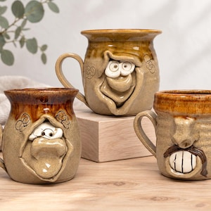 May include: Three handmade ceramic mugs with tan speckled glaze and brown rims. Each mug features a unique sculpted face with protruding noses, eyes, and mouths. The mugs have curved handles and are arranged on a light wooden surface.