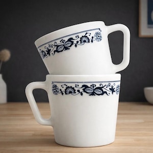 Puede incluir: Dos tazas de cerámica blanca apiladas. Cada taza tiene un asa grande y un diseño floral azul alrededor del borde. Las tazas están sobre una superficie de madera.