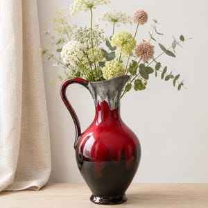 May include: A ceramic pitcher featuring a glossy deep red and black ombre glaze with a white drip effect around the rim and handle. The pitcher is filled with a bouquet of white, pale green, and dusty pink flowers with green eucalyptus foliage.