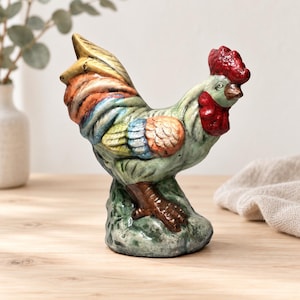Puede incluir: Una figura de gallo de cerámica con un cuerpo verde texturizado, una cresta y barbillas de color rojo brillante, y una cola con plumas multicolores en tonos naranja, azul y amarillo. El gallo se apoya sobre una base verde moteada con patas marrones.