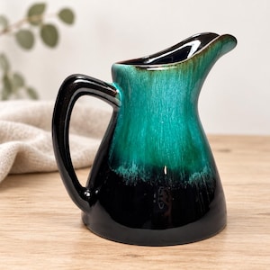 May include: A ceramic pitcher featuring a glossy glaze that transitions from a deep black at the base to a vibrant teal at the top. The vessel has a curved handle, a wide opening, and a prominent pouring spout with a smooth, reflective finish.