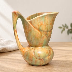 Puede incluir: Una jarra de cerámica con una base bulbosa y un pico ancho y acampanado. La superficie presenta un esmalte moteado en tonos mezclados de verde salvia, naranja cálido y crema, creando un efecto marmolado en todo el recipiente.