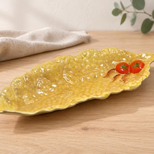 Puede incluir: Un plato de servicio de cerámica amarillo con forma de hoja y superficie texturizada. El plato presenta dos pequeñas decoraciones en relieve de tomates rojos con tallos verdes en el lado derecho, acentuadas por detalles de hojas marrones pintadas.