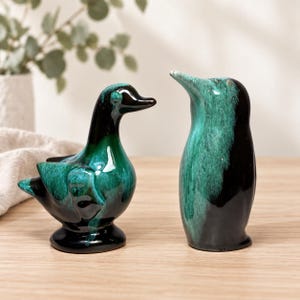 May include: Two ceramic figurines featuring a duck and a penguin, both finished with a glossy glaze in deep black and vibrant emerald green. The duck stands on a circular base, while the penguin has a smooth, upright, and elongated body shape.