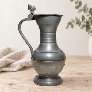 Puede incluir: Una jarra de metal gris con tapa, base bulbosa, cuello estrecho con estrías horizontales y un asa curva. La tapa está sujeta por una bisagra y rematada con un pequeño adorno decorativo en forma de un par de bellotas.