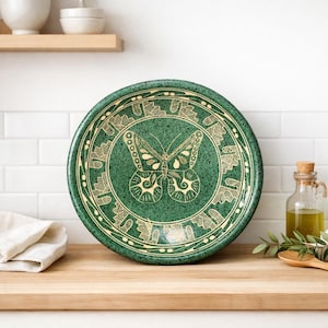 May include: A round, green ceramic plate featuring a cream-coloured butterfly design at its centre. The plate has a decorative border with leaf patterns. The plate is displayed on a wooden surface. The plate's diameter is approximately 25 cm.