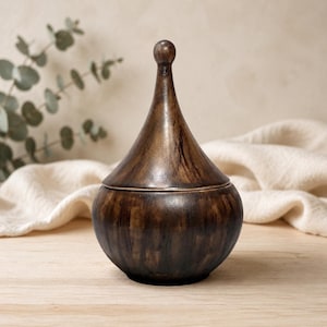 Puede incluir: Un recipiente redondeado de color marrón oscuro con una tapa a juego que presenta una parte superior alta, cónica y similar a un remate. La superficie del objeto muestra un acabado moteado similar a la veta de la madera con tonos marrones.