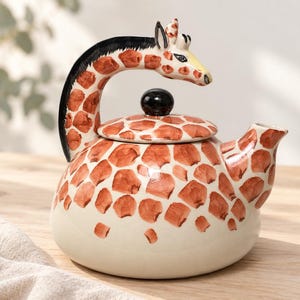May include: A ceramic teapot featuring a giraffe-inspired design with a cream-coloured base and brown patches. The handle is shaped like a giraffe neck and head with a black mane, and the lid includes a small, round, black knob for easy removal.