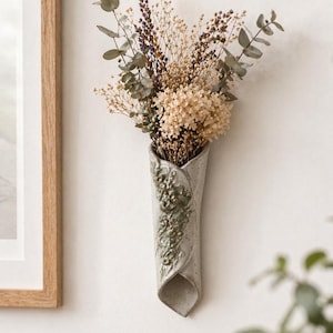 Puede incluir: Un jarrón de cerámica gris claro montado en la pared, con un ramo de flores secas. El jarrón tiene un diseño único y plegado con detalles florales en relieve. Las flores secas incluyen eucalipto, lavanda y otras flores beige y marrón, creando una estética rústica.
