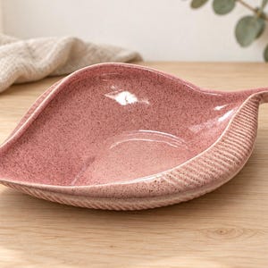 Peut inclure: Un bol en céramique en forme de feuille de couleur rose poudré avec une finition mouchetée. Le bol présente un motif texturé et nervuré le long du bord extérieur qui imite les veines d'une feuille, avec une surface intérieure lisse et brillante.