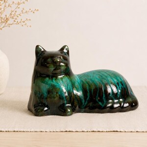 May include: A ceramic cat figurine in a reclining position, featuring a glossy glaze with deep black and vibrant teal green color variations that create a flowing, marbled effect across the smooth, rounded surface of the feline sculpture.
