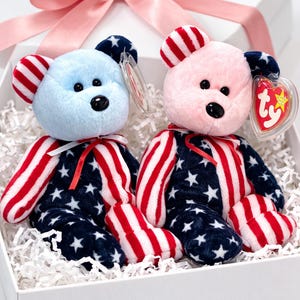 May include: Two plush teddy bears in a white gift box with a pink bow. One bear is light blue, the other is pink. Both have red and white striped ears and arms, and navy blue bodies with white stars. Each bear has a black nose and eyes.