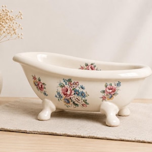 May include: A cream-colored ceramic bathtub-shaped dish featuring a floral pattern with pink, blue, and yellow flowers. The dish has four rounded feet and a smooth, glossy finish, designed as a decorative container for small items or toiletries.