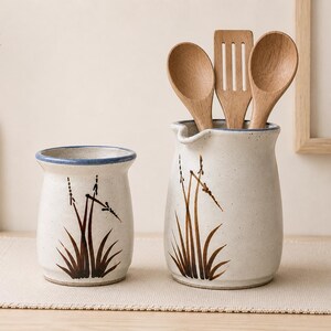 May include: Two ceramic kitchen utensil holders with a cream-coloured base and a blue rim. One holds wooden spoons and a spatula. Both feature a brown and black grass design. The holders sit on a beige placemat.