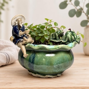 May include: A round, green glazed ceramic planter with a wavy rim. A figurine of an elderly person in a blue robe and straw hat sits on the edge. A second figurine of a green frog sits on the opposite rim. The planter contains a small green plant.