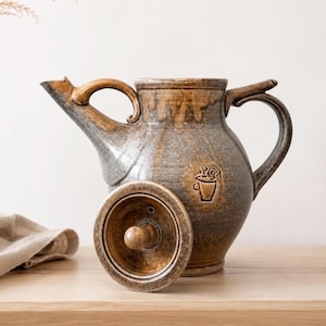 Può includere: Una caffettiera in ceramica fatta a mano, con coperchio, con una glassa marrone e grigia. La caffettiera ha un beccuccio, un manico e un emblema decorativo di una tazza di caffè fumante. Il coperchio è di lato.
