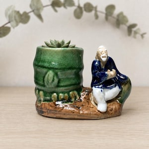 Può includere: Un vaso in ceramica composto da un contenitore verde testurizzato a forma di bambù con una piccola succulenta all'interno, accanto a una statuetta dipinta a mano di una persona anziana con barba bianca e tunica blu e bianca.