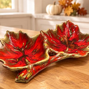 May include: A dual-section serving dish shaped like two connected maple leaves. The dish features a vibrant red glaze in the center of each leaf, transitioning to a metallic gold finish along the textured, raised edges and the central stem.