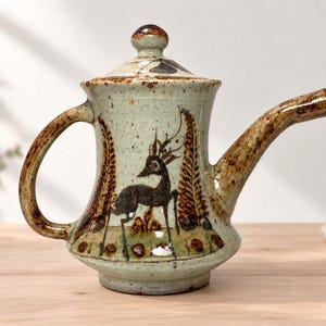 May include: A ceramic teapot with a light green speckled glaze, featuring a dark brown hand-painted deer illustration and fern-like patterns. The teapot includes a matching lid with a knob handle, a curved spout, and a sturdy side handle.