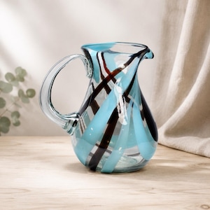May include: A glass jug featuring a rounded body and a curved handle. The clear glass is decorated with swirling, diagonal stripes in alternating shades of light blue and dark brown, creating a dynamic pattern across the entire surface.
