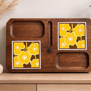 Puede incluir: Una bandeja de madera marrón oscuro con dos baldosas incrustadas que muestran un diseño floral amarillo sobre un fondo marrón. La bandeja tiene dos áreas empotradas y una ranura central con una pequeña perilla negra. Hay jarrones decorativos en el fondo.