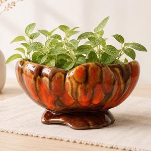 May include: A ceramic planter with a glossy, mottled orange and brown glaze, filled with green leafy plants. The planter has a scalloped rim and sits on a decorative base. A white vase with dried flowers is in the background.