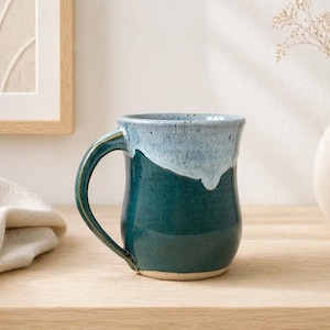 Può includere: Una tazza in ceramica fatta a mano con base verde acqua e parte superiore azzurra. La tazza ha un manico curvo e una finitura lucida. La tazza è appoggiata su una superficie di legno.