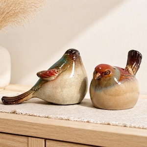 May include: Two ceramic bird figurines. One bird is elongated with a brown, teal, and red color scheme. The other bird is rounder, with a brown and tan color scheme. Both are placed on a neutral-colored surface.