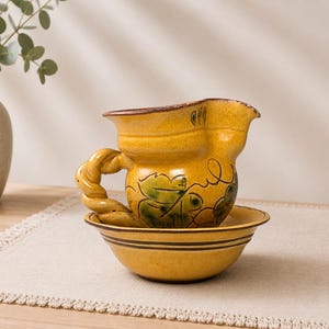 Puede incluir: Un juego de jarra y cuenco de cerámica amarilla con un motivo de hojas verdes pintado a mano en la jarra. La jarra tiene un asa trenzada y un pico ancho, mientras que el cuenco está decorado con bandas horizontales de color marrón oscuro.