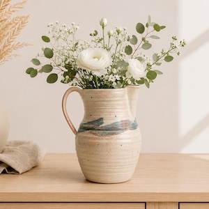Peut inclure: Un pichet en céramique avec une finition crème mouchetée et une anse unique, présentant une bande horizontale de glaçure bleue et brune au centre. Le pichet est rempli d'un bouquet de renoncules blanches, de gypsophile et de feuilles d'eucalyptus.