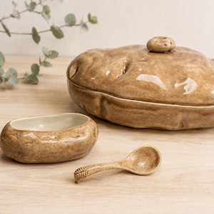 May include: A ceramic set shaped like potatoes, including a large lidded container, a small open bowl with a cream-colored interior, and a matching spoon. Each piece features a glossy, textured brown finish with realistic indentations.