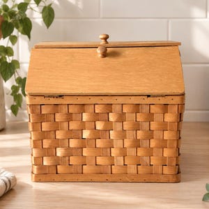 May include: A rectangular storage basket with a woven light brown wood base and a solid wood hinged lid. The lid features two small round wooden knobs on the front edge. The basket has a structured design with a flat top and vertical sides.