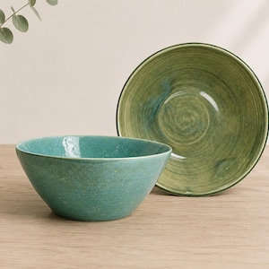 May include: Two ceramic bowls with a smooth, rounded shape. One bowl is a vibrant turquoise colour with a subtle speckled texture, while the other is a larger, deeper green bowl featuring visible circular brushstroke patterns on its interior surface.