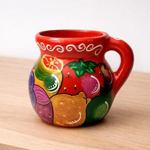 May include: A hand-painted ceramic mug with a red exterior and a handle. The mug is decorated with colorful images of fruits and vegetables, including tomatoes, avocados, and onions. The rim features a white swirl design.
