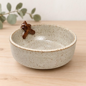Peut inclure: Un bol en céramique rond avec une finition gris clair mouchetée et une texture lisse. Une petite figurine d'abeille tridimensionnelle brun foncé est fixée sur le bord intérieur du bol, offrant un contraste net avec la surface claire mouchetée.