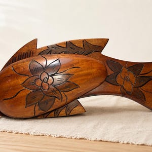 Puede incluir: Una escultura de madera marrón con forma de pez que presenta patrones florales tallados a mano en el cuerpo y la cola. La madera tiene un acabado pulido con detalles en marrón oscuro que resaltan los pétalos y las hojas grabadas.