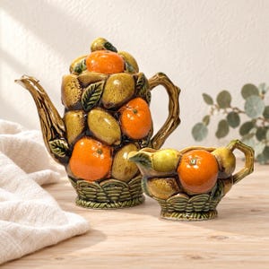 Puede incluir: Un juego de tetera y lechera de cerámica con un diseño en relieve tridimensional de naranjas y limones. Las piezas tienen un acabado brillante con detalles de hojas verdes y una base de cestería, con asas en forma de ramas de árbol.
