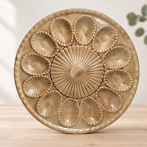 May include: A round, tan-colored ceramic oyster plate featuring a central circular section with radiating ridges, surrounded by twelve individual oval-shaped indentations, each with a scalloped edge and a glossy, glazed finish.