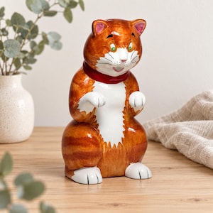 May include: A ceramic cat figurine featuring a vibrant orange tabby coat with white markings on the chest, paws, and face. The cat has green eyes, a small pink nose, and a dark red collar around its neck, depicted in a standing, upright posture.