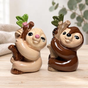 Peut inclure: Deux figurines de paresseux en céramique, l'une beige clair et l'autre marron foncé, agrippées à une branche d'arbre marron texturée avec des feuilles vertes. Les deux paresseux ont une finition brillante, des yeux noirs et de petits nez.