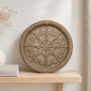 May include: A round, beige decorative tray with an intricate heart and leaf design. The tray is made of a textured material and has a raised edge. It sits on a light wood shelf with a vase and a book.