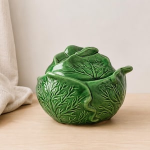Puede incluir: Un cuenco de cerámica verde brillante con forma de cabeza de repollo y tapa a juego. El exterior presenta intrincadas texturas de nervaduras de hojas en relieve y bordes ondulados, imitando el aspecto natural de un repollo en verde vibrante.