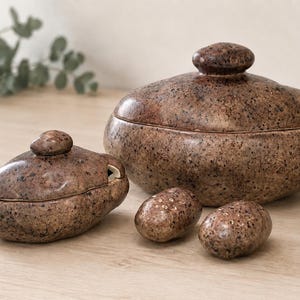 May include: A set of brown speckled ceramic kitchenware consisting of a large lidded bowl, a smaller lidded container with a side opening, and two small oval shakers with perforated tops. Each piece features a smooth, glossy, stone-like finish.