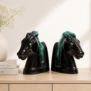 May include: Pair of black ceramic horse head bookends with teal accents. The bookends have a glossy finish and are placed on a light wood surface. A vase with white flowers and two books are also in the image.