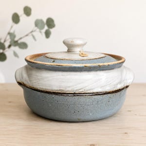Puede incluir: Una cazuela de cerámica redonda con tapa, que presenta un diseño bicolor con una base de color azul grisáceo moteado y una sección superior blanca texturizada. La tapa tiene un pomo central y una pequeña etiqueta color canela. Dos asas laterales.