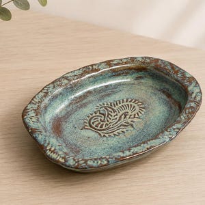 Peut inclure: Un plat ovale en céramique avec un émail moucheté bleu sarcelle et brun. Le bord présente une texture festonnée, et le centre arbore un motif estampé complexe rappelant un cachemire brun foncé, contrastant avec la base bleu-vert plus claire.